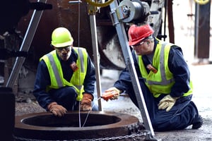 Two industrial workers kneel and use a gas monitor with a sampling pump to read gas levels in a confined space