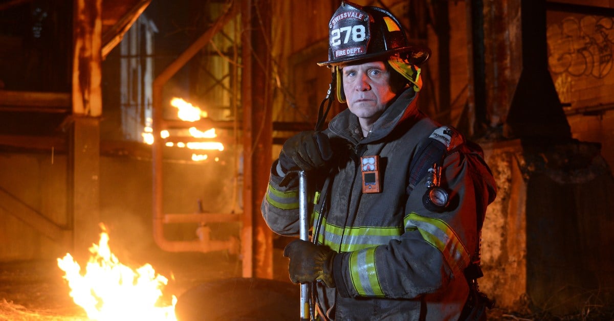 A fire fighter stares into the camera. Behind him, flames spread around a structure.