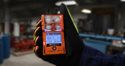 gloved hand holds a personal gas monitor with a screen that shows an alarm for hydrogen sulfide