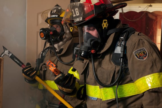 Two fire fighters wearing personal protective equipment use a personal gas monitor to read the atmosphere in a house.