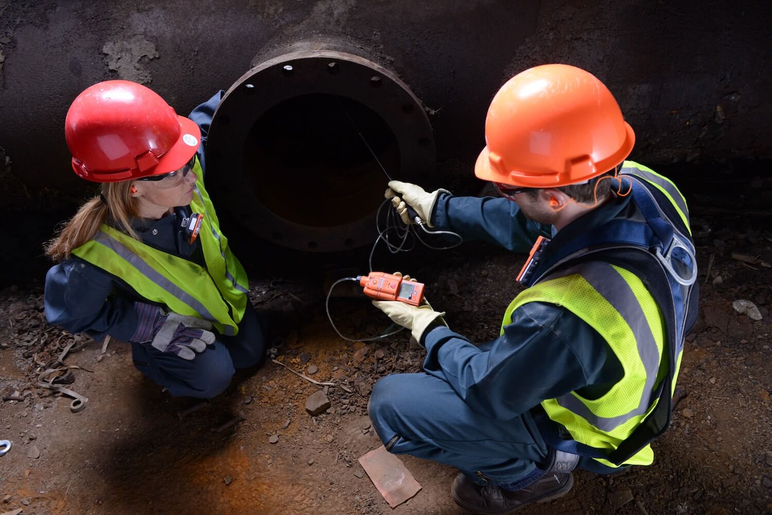 Two industrial workers kneel and use a gas monitor with a sampling pump to read gas levels in a confined space