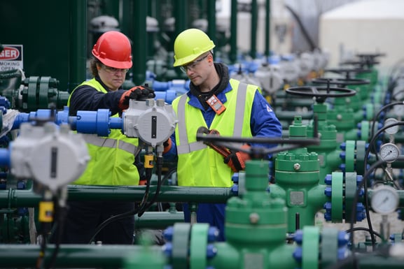 Two industrial workers wearing personal protective equipment and gas monitors inspect a piece of equipment