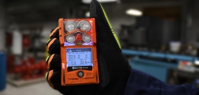 Hand holds up a Ventis Pro5 gas monitor. The screen shows an alert for high H2S