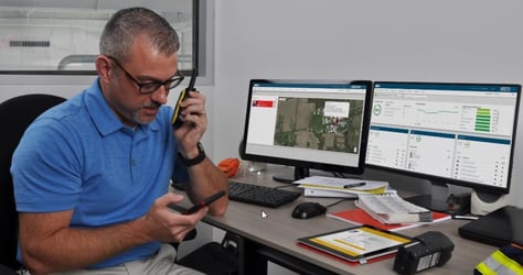 safety manager in an office looks at notification on his phone while making a call on a walkie-talkie. On his computer screens, there is an alert showing a gas exposure to one of the employees