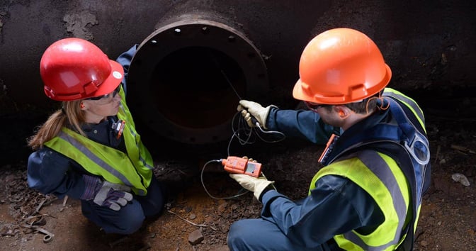 Two industrial workers kneel and use a gas monitor with a sampling pump to read gas levels in a confined space