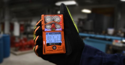 gloved hand holds up a personal gas monitor displaying an alarm for hydrogen sulfide