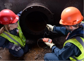 Two industrial workers kneel and use a gas monitor with a sampling pump to read gas levels in a confined space