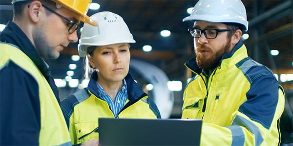 three industrial workers in hardhats look at a screen while the manager directs their attention to something. 