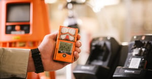 In the background are docking stations and an area monitor. In the foreground, a hand holds out an orange personal gas monitor.