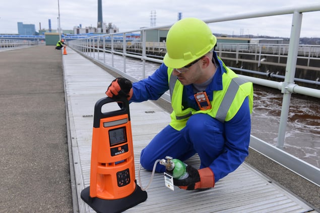 worker on a bridge wearing PPE and gas monitor kneels to look at screen on an area monitor