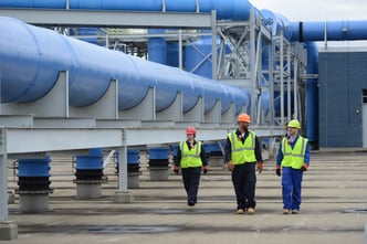 three workers wearing personal gas monitors walk along a pipeline